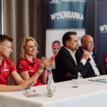 Panel of five people seated at a long table during a formal event; man in a suit speaks into a microphone while others applaud, red shirts worn by some panelists, WYSOWANKA banners in the background.