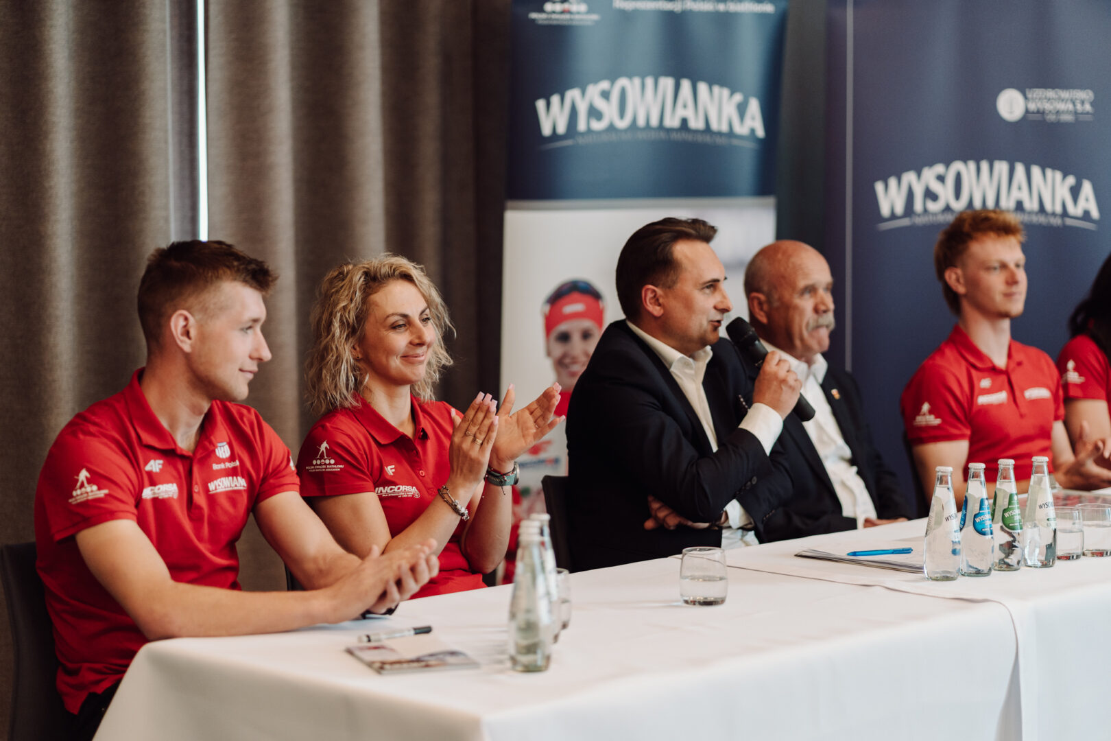 Panel of five people seated at a long table during a formal event; man in a suit speaks into a microphone while others applaud, red shirts worn by some panelists, WYSOWANKA banners in the background.