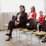 Woman in a brown blazer sits in the front row with legs crossed, attention forward; two people in the back are seated in a bright room.