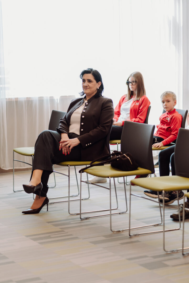 Woman in a brown blazer sits in the front row with legs crossed, attention forward; two people in the back are seated in a bright room.