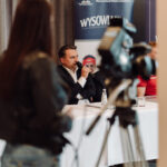 Man in a suit speaks into a microphone at a panel table during a press conference, with a media crew and a banner in the background.