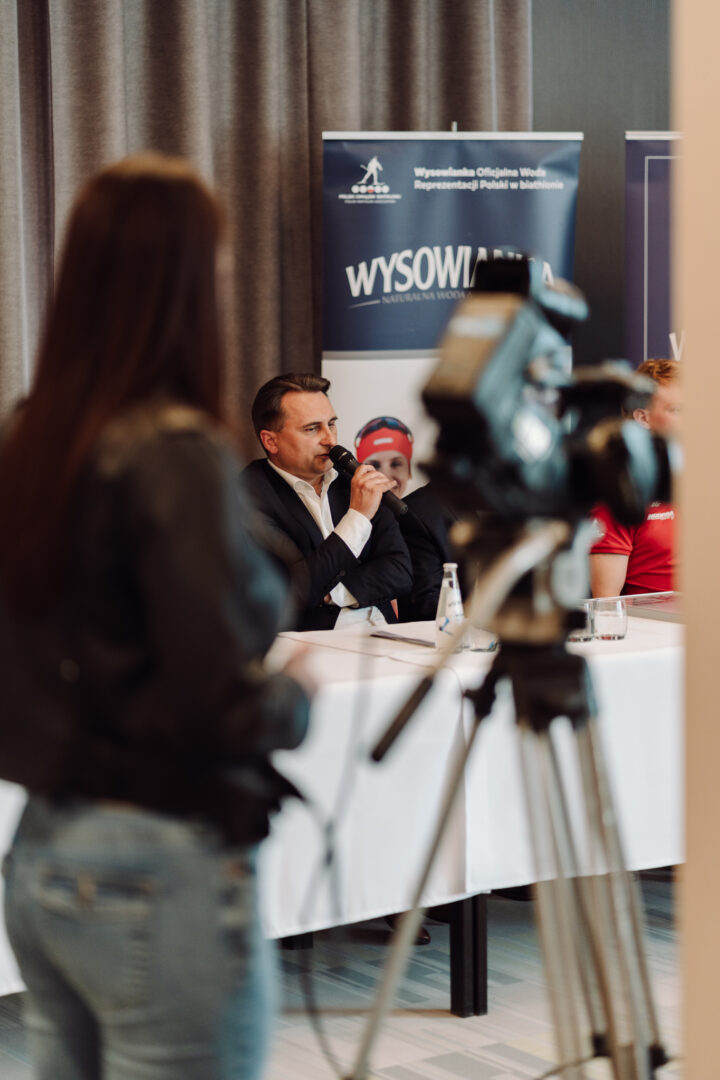 Man in a suit speaks into a microphone at a panel table during a press conference, with a media crew and a banner in the background.
