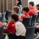 Group of young students in red and white shirts seated in a classroom, listening to a speaker.