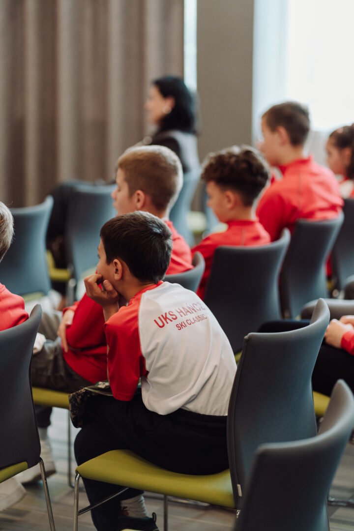 Group of young students in red and white shirts seated in a classroom, listening to a speaker.