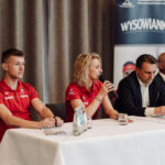 Panel of five people seated at a long table during a press event; two in red polo shirts, two men in suits, microphones and water bottles on the table.