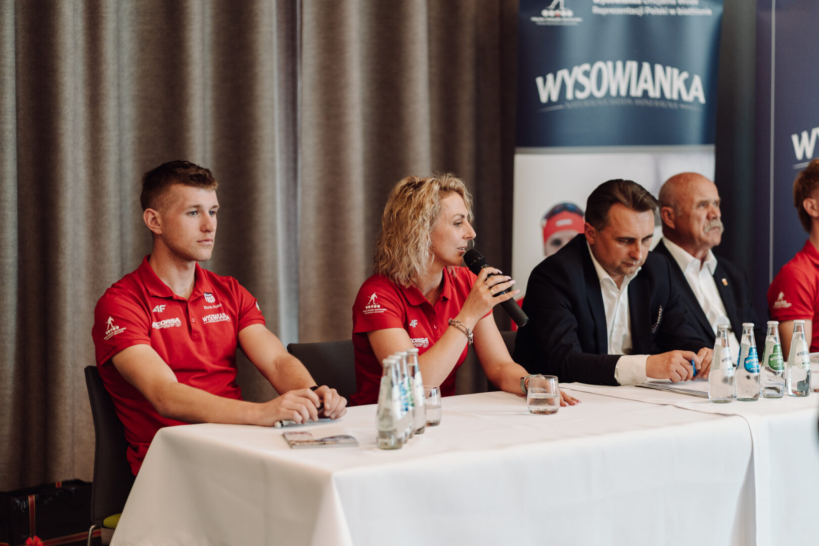 Panel of five people seated at a long table during a press event; two in red polo shirts, two men in suits, microphones and water bottles on the table.