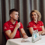 Two panelists in red polo shirts sit at a table, the man speaking into a handheld microphone while the woman listens to him. A row of water bottles and glasses sit on the table.