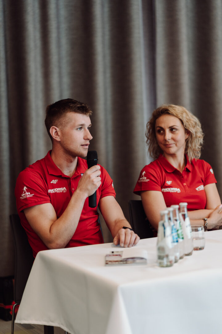 Two panelists in red polo shirts sit at a table, the man speaking into a handheld microphone while the woman listens to him. A row of water bottles and glasses sit on the table.