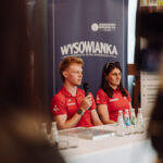 Two presenters in red polo shirts sit at a panel table with a microphone, in front of a blue banner that reads WYSOWIANKA.