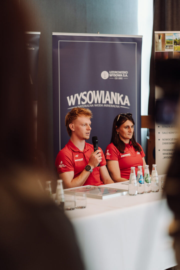 Two presenters in red polo shirts sit at a panel table with a microphone, in front of a blue banner that reads WYSOWIANKA.