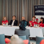 Panel of six people seated at a long white-table conference, with two banners reading 'WYSOWIANKA' in the background and several water bottles on the table (informational event).