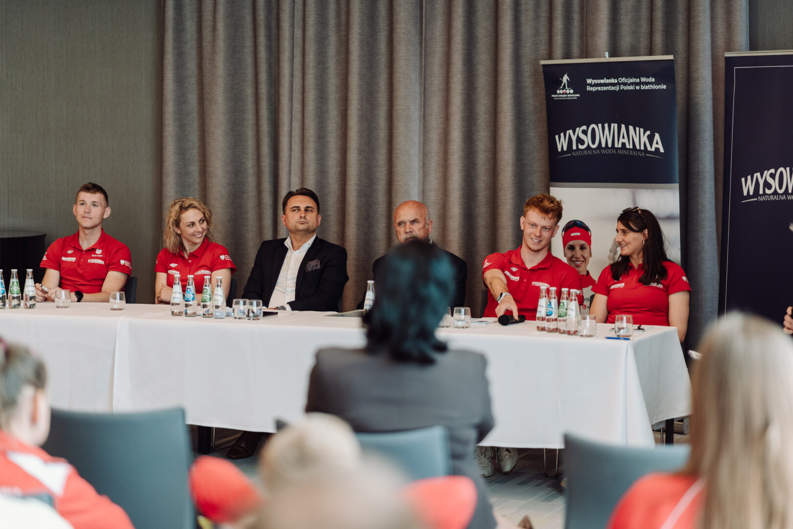 Panel of six people seated at a long white-table conference, with two banners reading 'WYSOWIANKA' in the background and several water bottles on the table (informational event).