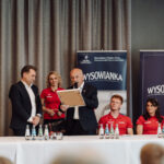 Panel discussion at a conference: a suited man presents a framed award while two people in red shirts and another suited man look on, with banners reading 'WYSOWANKA' in the background.