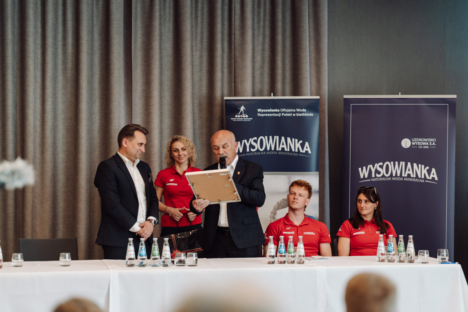 Panel discussion at a conference: a suited man presents a framed award while two people in red shirts and another suited man look on, with banners reading 'WYSOWANKA' in the background.