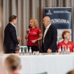 Woman in a red polo speaks into a microphone at a panel, flanked by two men in suits; a sponsor banner and water bottles sit on the table behind them.