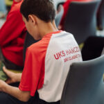 Young athlete in a red and white UKS ski team jersey sits in a chair, looking at a camouflage cap in their hands.