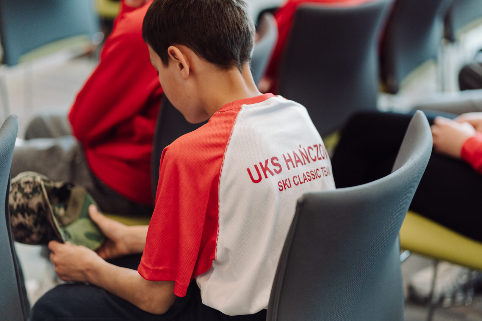 Young athlete in a red and white UKS ski team jersey sits in a chair, looking at a camouflage cap in their hands.