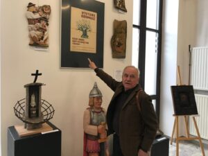 Older man in a brown jacket points to a poster on a gallery wall amid wooden sculptures and display cases.