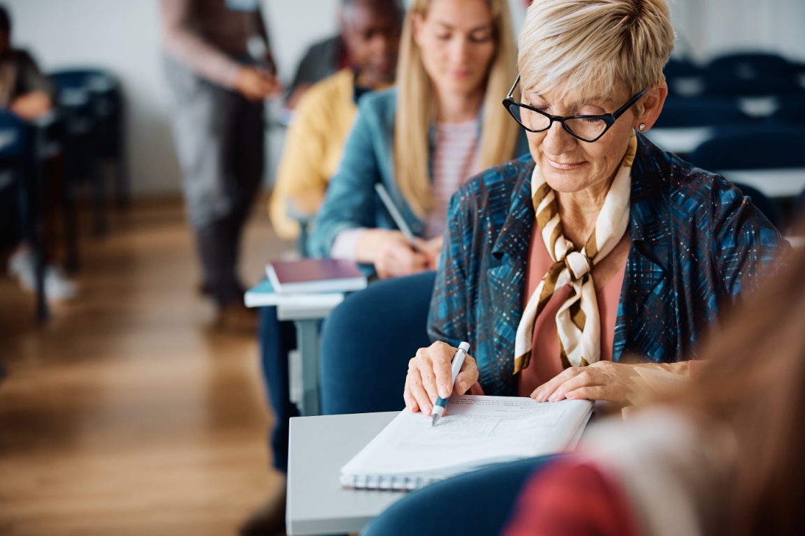 Older woman with glasses writing in a notebook at a classroom desk, others studying in the background.
