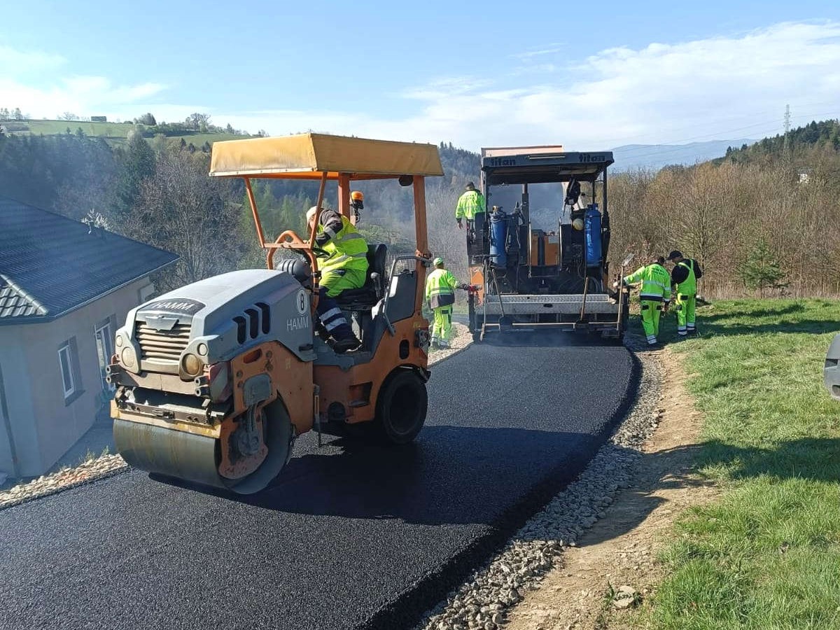 Road construction crew laying fresh asphalt on a rural hillside road with a roller compactor and asphalt paver in use