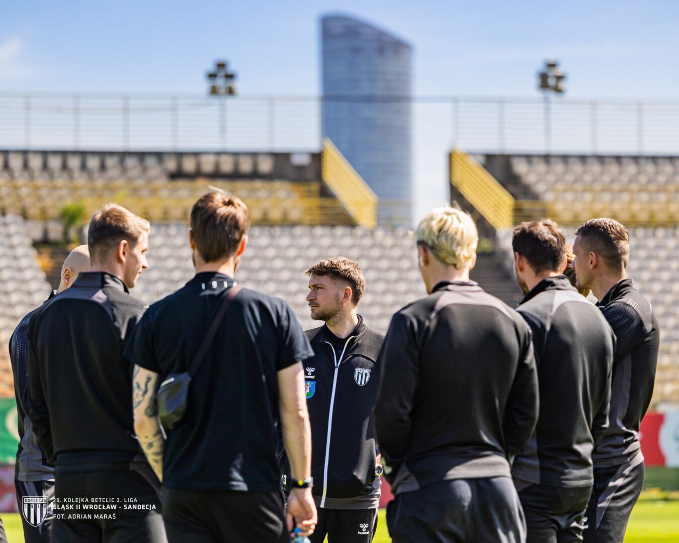 Coach addressing a group of football players in a circle on a sunny training field outside a stadium.