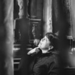 Black-and-white photo of a person in a dark jacket looking up in a dim, vintage room with heavy curtains and wooden furnishings.