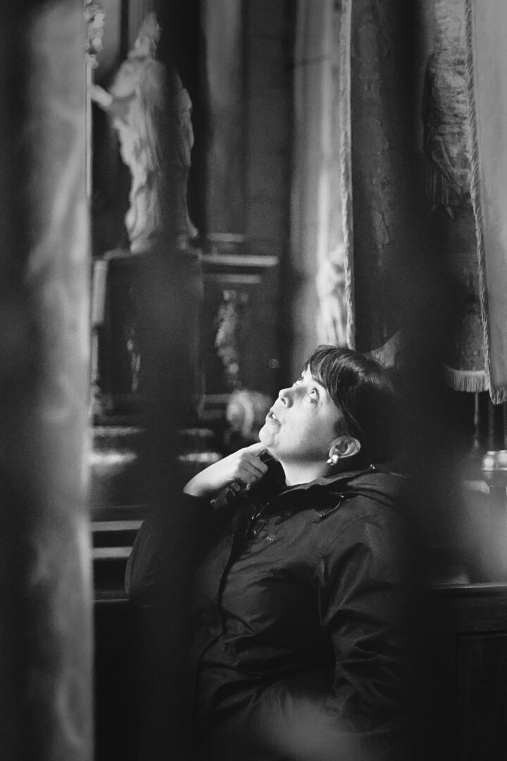 Black-and-white photo of a person in a dark jacket looking up in a dim, vintage room with heavy curtains and wooden furnishings.