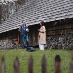 Man and woman conversing beside a rustic log cabin with a large woodpile in a grassy yard.