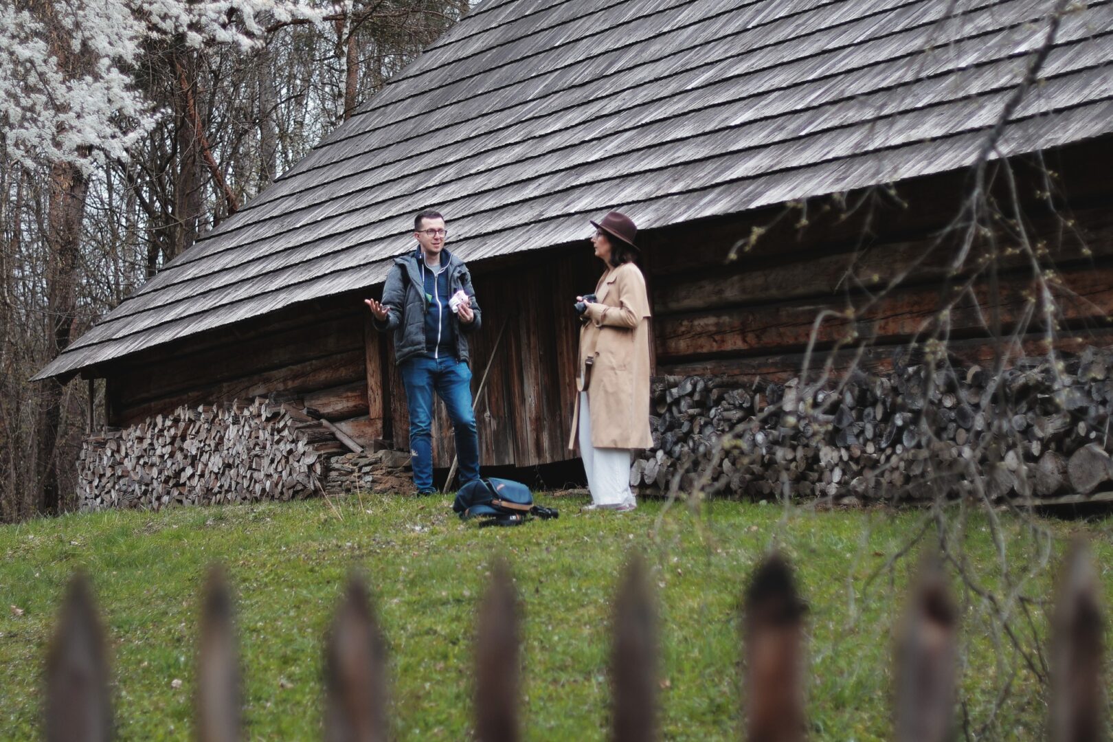 Man and woman conversing beside a rustic log cabin with a large woodpile in a grassy yard.