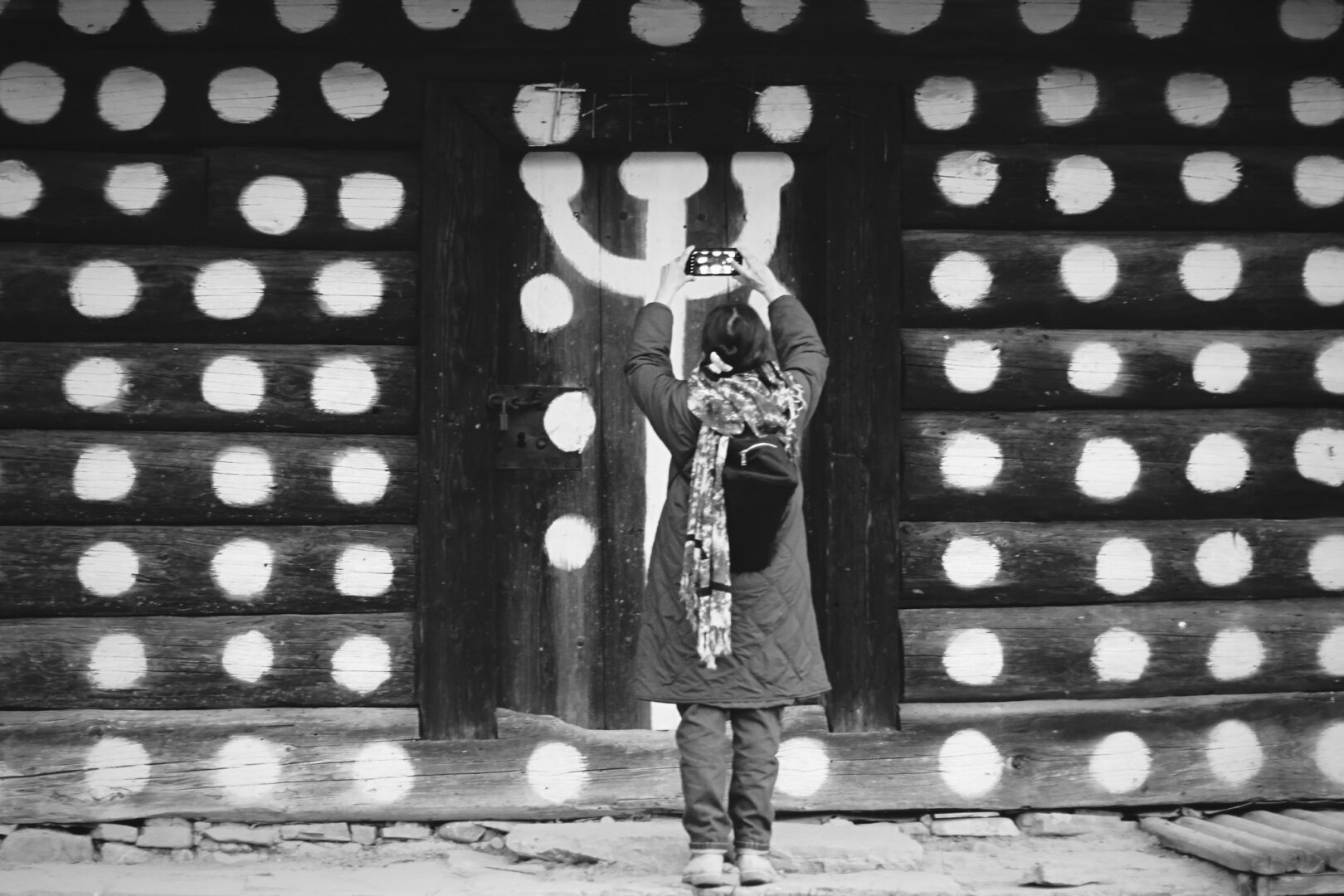 Person standing with a backpack facing a large wooden door, taking a photo with a smartphone of white circular patterns on the door exterior.
