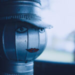 Close-up of a weathered metal lantern shaped as a face with a cap and red lips, front view in blue-tinted light.