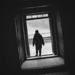 Silhouette of a person standing in a doorway, looking out at a bright yard outside.