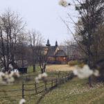 Wooden church with two domed towers in a rural village, framed by bare trees and a split-rail fence in the foreground.