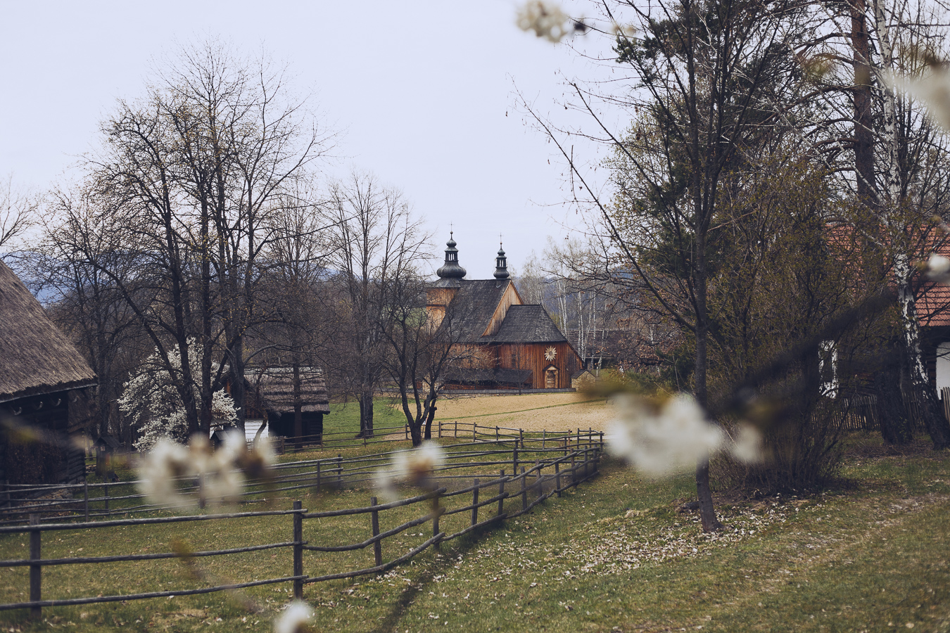 Wooden church with two domed towers in a rural village, framed by bare trees and a split-rail fence in the foreground.