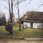 Thatched-roof cottage with white walls along a grassy path, next to a wooden split-rail fence and a wicker basket on the gate post.