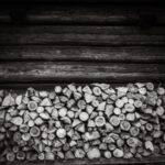 Stack of chopped firewood piled against a weathered log wall in black and white.