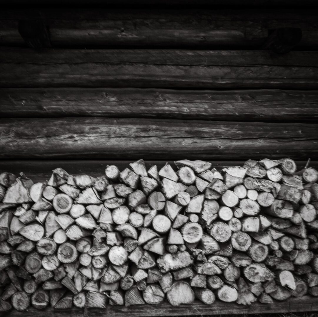 Stack of chopped firewood piled against a weathered log wall in black and white.