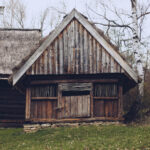 Weathered wooden barn with a steep gabled roof and thatched section, set in a grassy yard with leafless trees in the background