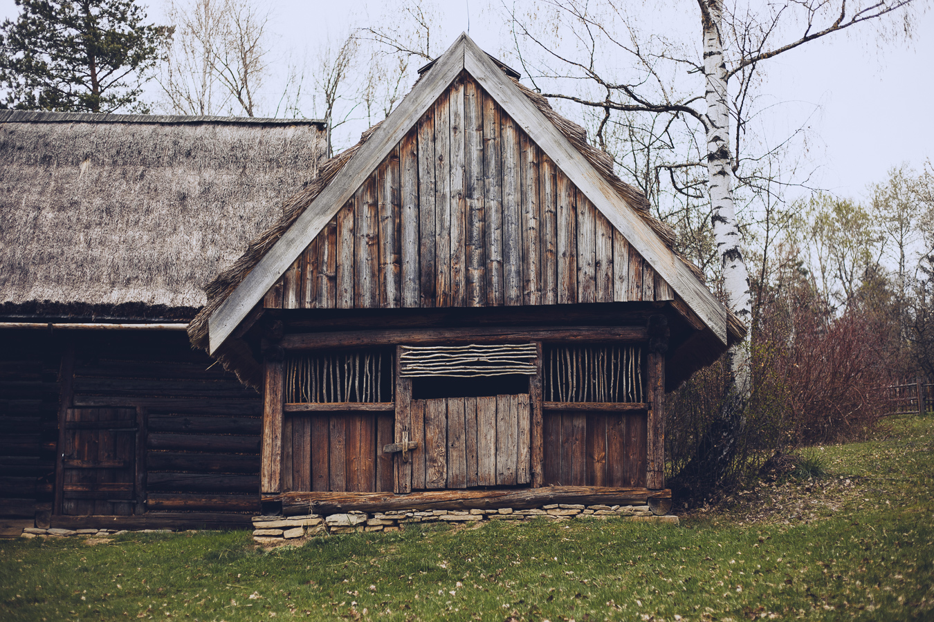 Weathered wooden barn with a steep gabled roof and thatched section, set in a grassy yard with leafless trees in the background