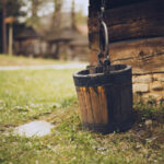 Wooden bucket hanging from a metal chain beside a rustic log cabin in a grassy yard.