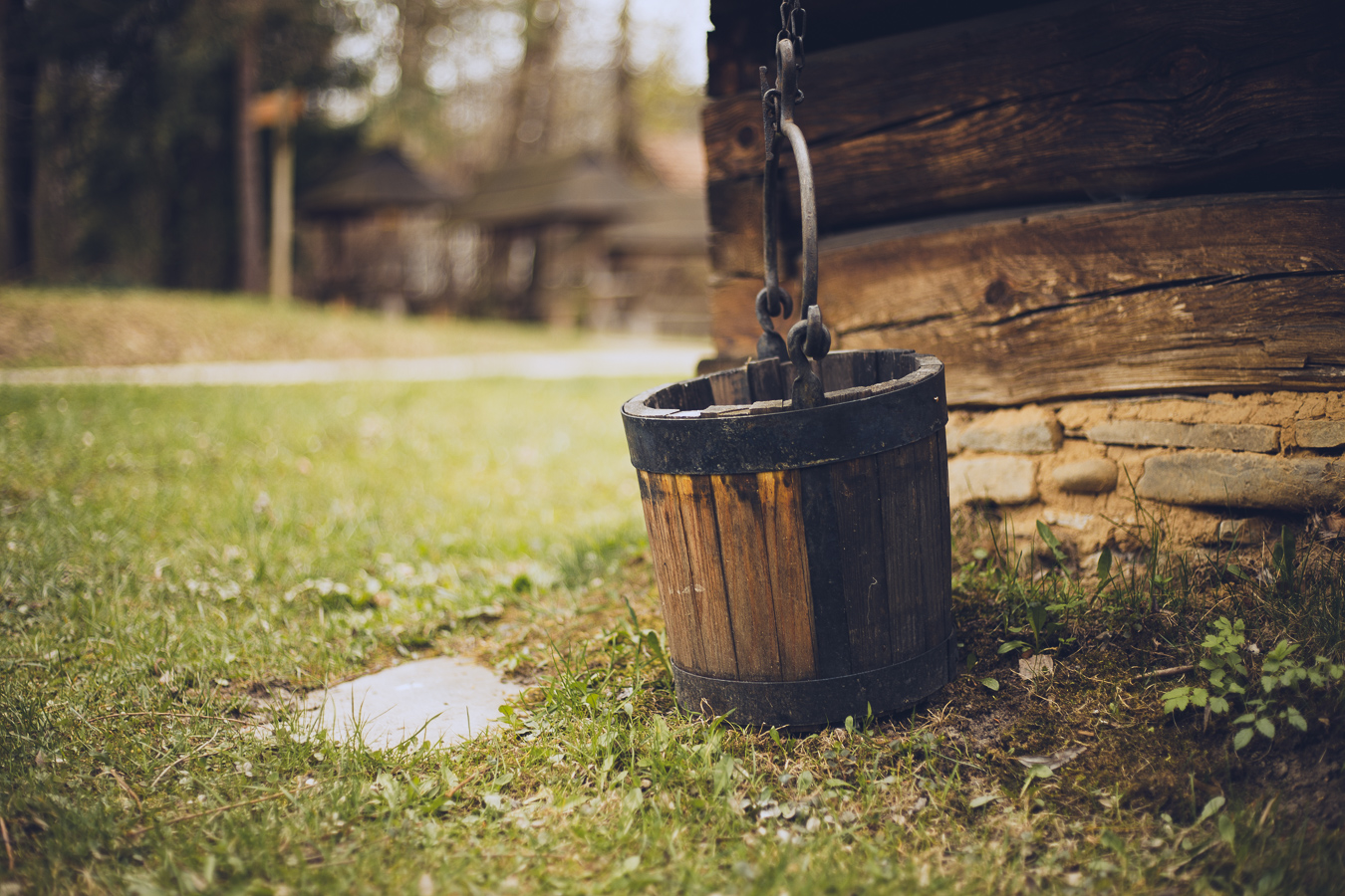 Wooden bucket hanging from a metal chain beside a rustic log cabin in a grassy yard.