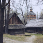 Cluster of weathered wooden cabins among leafless trees; a wooden church tower visible in the distance.