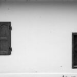 White exterior wall with a closed dark wooden shutter on the left and a small dark wooden door with diagonal panels on the right; a round fire alarm sign reading 'POŽAR' is at the top left.