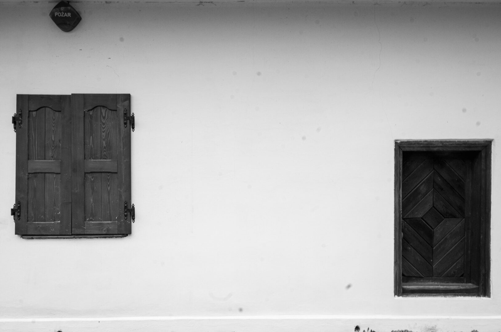 White exterior wall with a closed dark wooden shutter on the left and a small dark wooden door with diagonal panels on the right; a round fire alarm sign reading 'POŽAR' is at the top left.