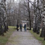Three people conversing on a gravel path lined with birch trees in a park, with a small wooden shed in the background.