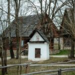Small white shrine or well building with a wooden barred door, set in a yard with bare trees and wooden houses behind; a rustic fence in the foreground.