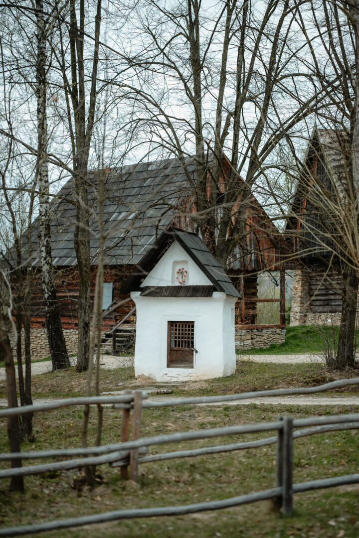 Small white shrine or well building with a wooden barred door, set in a yard with bare trees and wooden houses behind; a rustic fence in the foreground.