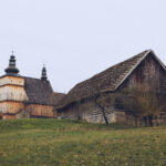 Old wooden church with twin onion-domed towers beside a weathered log barn in a rural landscape.