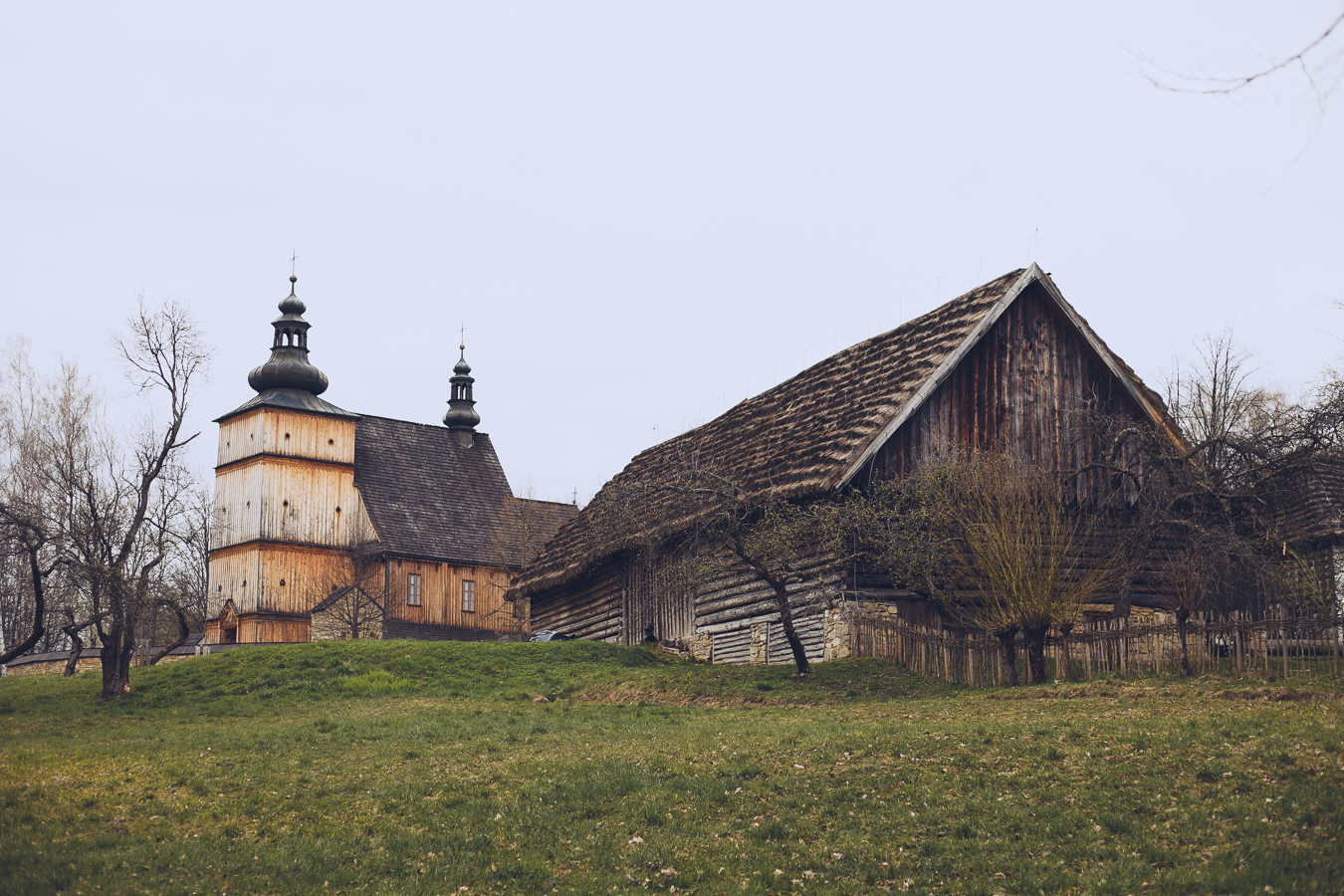 Old wooden church with twin onion-domed towers beside a weathered log barn in a rural landscape.