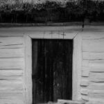 Weathered wooden door in a whitewashed log cabin with a thatched roof, small crosses above the doorway.
